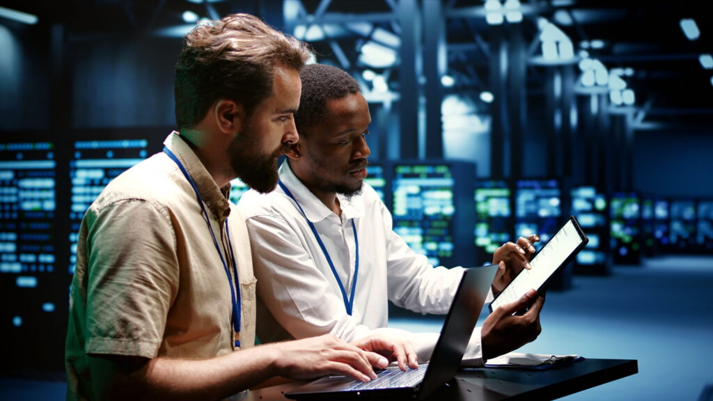 Two computer science professionals standing in a data center with a laptop