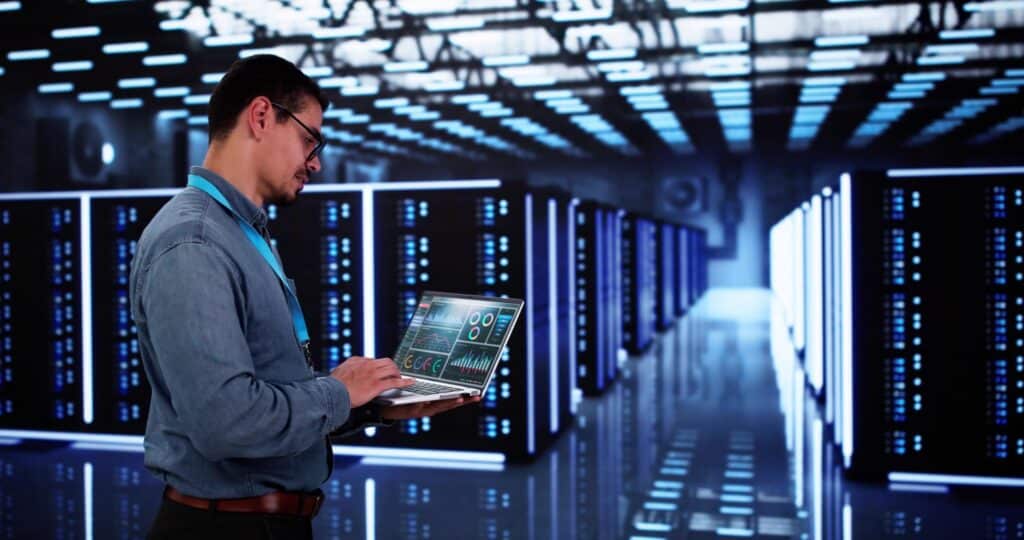 Data analyst standing in a data center with a laptop, working an AI job with a bachelor's degree